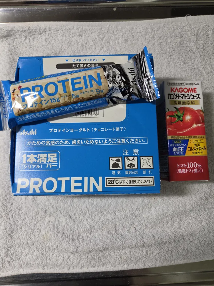 アサヒ1本満足バー プロテインの箱と、開封した1本のチョコクランチ断面。右にはおなじみの大きさ比較用トマトジュース